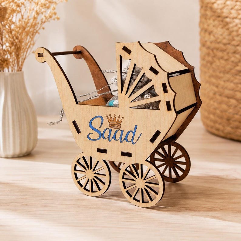 A laser-cut wooden toy baby stroller with the name Saad and a small crown printed in blue on the side, sitting on a wooden surface with a white vase of dried flowers in the background.