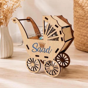 A wooden laser-cut baby carriage model with four spoked wheels sits on a light wooden surface, featuring the blue engraved name Saad below a small crown.