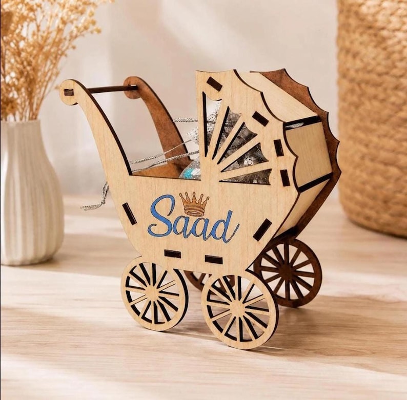 A wooden laser-cut baby carriage model with four spoked wheels sits on a light wooden surface, featuring the blue engraved name Saad below a small crown.