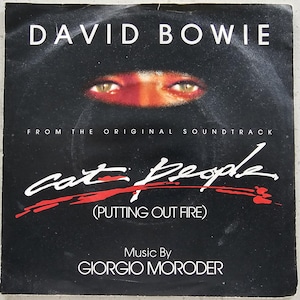Puede incluir: Un disco de vinilo negro y blanco de 7 pulgadas con un gráfico rojo y blanco del texto "cat people (putting out fire)" y el nombre del artista "DAVID BOWIE". El disco es de la banda sonora original de la película "Cat People".