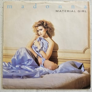 Puede incluir: Una mujer en sujetador blanco y una sábana de satén azul claro posa en una cama. El texto "Madonna" y "Material Girl" está impreso en el fondo blanco.