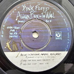 May include: A black 45 RPM vinyl record with a label featuring the text Pink Floyd Another Brick in the Wall Part II. The label has a blue and white background with a cartoon character, the Harvest logo, and production credits from 1979.