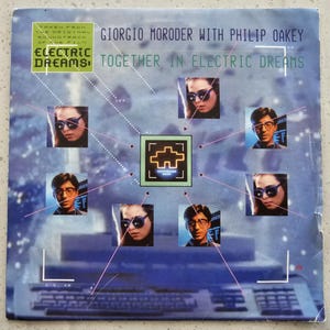 May include: Album cover for "Together in Electric Dreams" by Giorgio Moroder with Philip Oakey. The cover features a blue background with images of people wearing sunglasses, connected by pink lines. Text includes "Electric Dreams" and artist names.