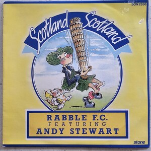 May include: A vintage vinyl record featuring a cartoon illustration of a Scottish figure struggling to hold up the Leaning Tower of Pisa. The record has the text "Scotland" and "RABBLE F.C. FEATURING ANDY STEWART" on a yellow background.