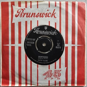May include: Vintage 45 RPM record by Patsy Cline, titled "Heartaches." The record is black with white text and a center hole. It is housed in a red and white striped sleeve with the "Brunswick" logo.