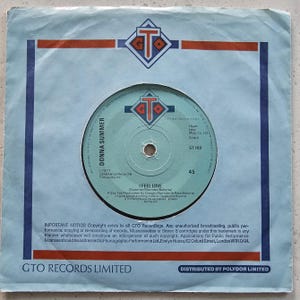 Può includere: Un disco in vinile vintage di Donna Summer, intitolato "I Feel Love", è esposto nella sua custodia originale. L'etichetta del disco è azzurra con il logo GTO Records. La custodia ha un bordo blu con accenti rossi e arancioni.