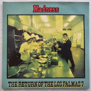 May include: Album cover for Madness' "The Return of the Los Palmas 7". The image depicts a kitchen with chefs in white uniforms and tall hats, and a group of men in suits. The band's name is in red at the top, and the album title is at the bottom.