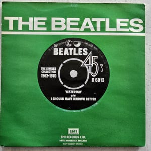 Può includere: Una custodia per dischi in vinile verde con un'etichetta in bianco e nero. L'etichetta presenta il logo dei Beatles, il titolo "The Beatles 45's", il titolo della canzone "Yesterday" e il titolo della canzone "I Should Have Known Better". L'etichetta include anche il testo "The Singles Collection 1962-1970" e "R 6013". La custodia include anche il testo "EMI Records Ltd., Hayes Middlesex England, Made in Great Britain."