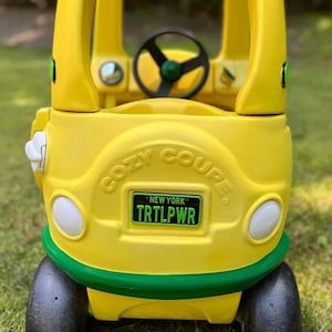 May include: A yellow and green Cozy Coupe toy car with a steering wheel and a license plate that reads "NEW YORK TRTLPWR".