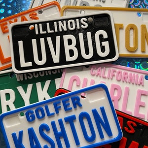 Puede incluir: Una colecci&oacute;n de placas de matr&iacute;cula personalizadas de colores con diferentes nombres y estados. Las placas son de pl&aacute;stico y tienen un efecto 3D. Los nombres en las placas incluyen "LUVBUG", "KASHTON", "RYK" y "CHARLIE".
