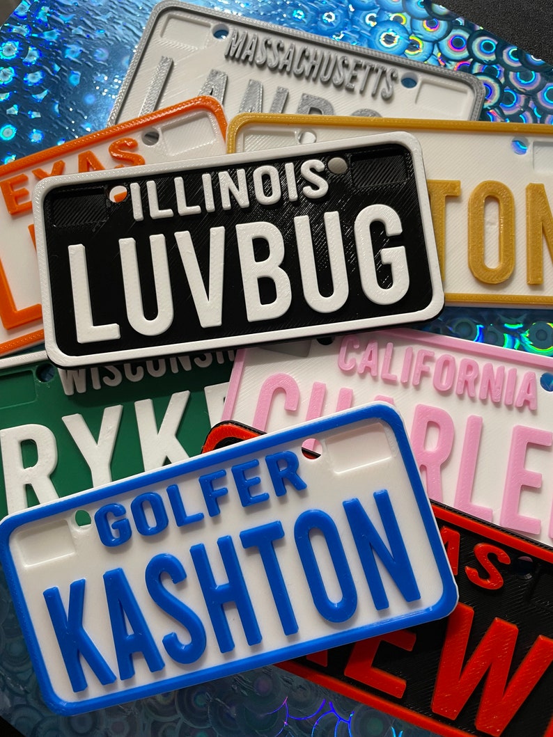 Puede incluir: Una colecci&oacute;n de placas de matr&iacute;cula personalizadas de colores con diferentes nombres y estados. Las placas son de pl&aacute;stico y tienen un efecto 3D. Los nombres en las placas incluyen "LUVBUG", "KASHTON", "RYK" y "CHARLIE".
