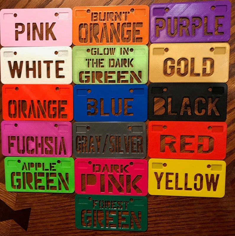 Puede incluir: Una colecci&oacute;n de 15 peque&ntilde;as etiquetas de pl&aacute;stico con forma de matr&iacute;cula, en varios colores, cada una con un nombre de color diferente impreso en ella. Los colores incluyen rosa, naranja, morado, blanco, verde, dorado, azul, negro, fucsia, gris/plata, rojo, verde manzana, rosa oscuro, amarillo y verde bosque.