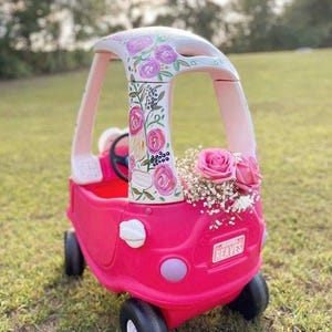 May include: A bright pink plastic toy car featuring a white roof painted with pink and green floral designs. The front of the car is decorated with a cluster of pink roses and white baby's breath flowers. A license plate reads REAVES.