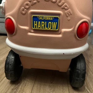 Puede incluir: Un coche de juguete de pl&aacute;stico rosa con un parachoques blanco y ruedas negras. El coche tiene una matr&iacute;cula que dice "CALIFORNIA HARLOW".