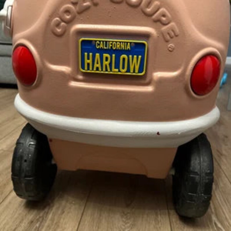 Puede incluir: Un coche de juguete de pl&aacute;stico rosa con un parachoques blanco y ruedas negras. El coche tiene una matr&iacute;cula que dice "CALIFORNIA HARLOW".