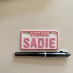 Puede incluir: Placa de matr&iacute;cula de pl&aacute;stico rosa con la palabra "VIRGINIA" y el nombre "SADIE" en letras blancas.