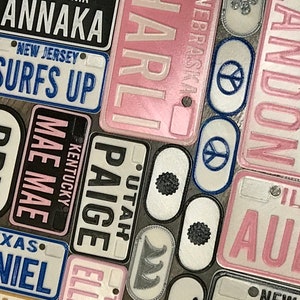 Puede incluir: Una colecci&oacute;n de placas de matr&iacute;cula personalizadas con varios nombres y estados. Las placas son de metal y tienen un fondo blanco con letras negras. Algunas de las placas tienen un fondo rosa con letras blancas. Las placas est&aacute;n dispuestas en un orden aleatorio.