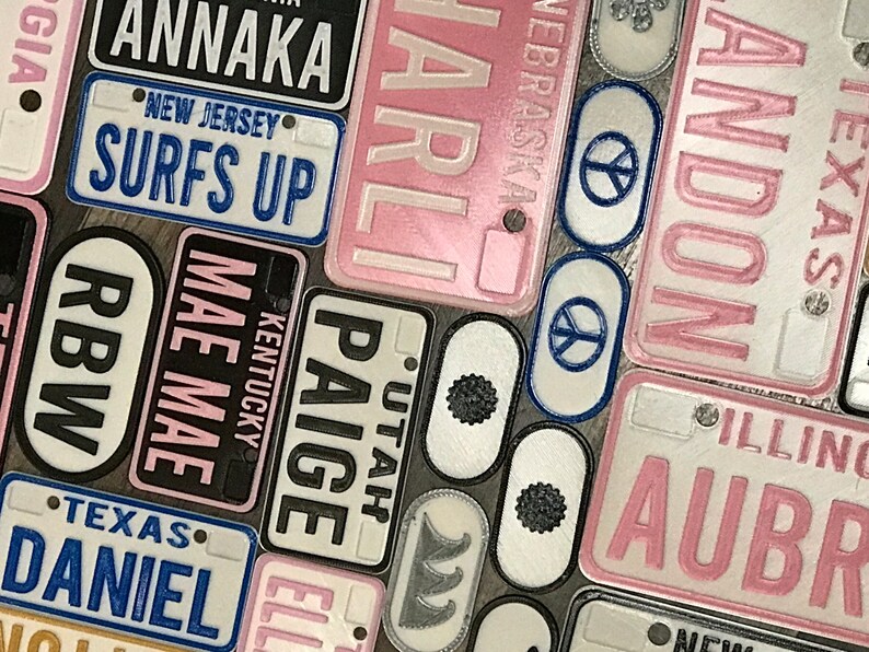 Puede incluir: Una colecci&oacute;n de placas de matr&iacute;cula personalizadas con varios nombres y estados. Las placas son de metal y tienen un fondo blanco con letras negras. Algunas de las placas tienen un fondo rosa con letras blancas. Las placas est&aacute;n dispuestas en un orden aleatorio.