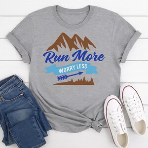 Puede incluir: Camiseta gris con la frase "Run More Worry Less" en azul y un gr&aacute;fico de monta&ntilde;a. El dise&ntilde;o incluye una pancarta azul, una flecha y una silueta de bosque marr&oacute;n. La camiseta se muestra con vaqueros azules y zapatillas blancas.