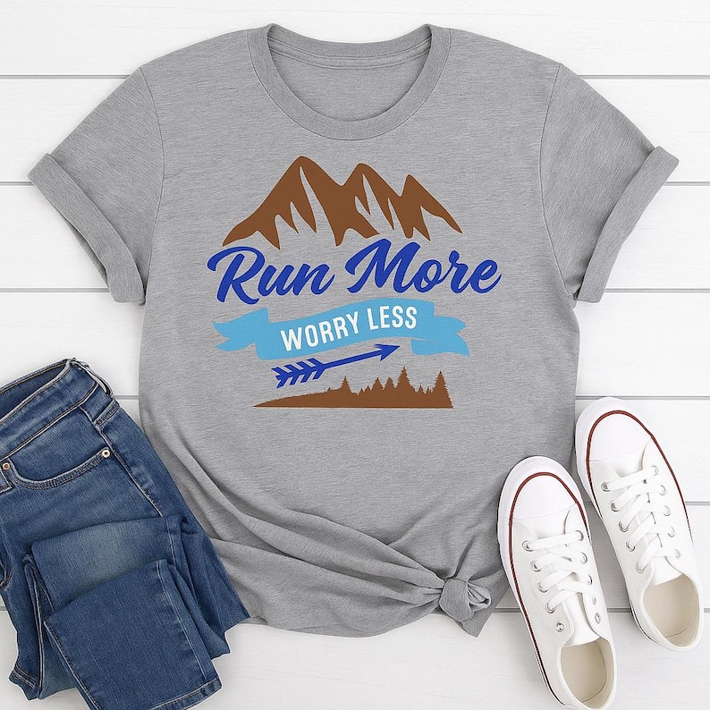 Puede incluir: Camiseta gris con la frase "Run More Worry Less" en azul y un gr&aacute;fico de monta&ntilde;a. El dise&ntilde;o incluye una pancarta azul, una flecha y una silueta de bosque marr&oacute;n. La camiseta se muestra con vaqueros azules y zapatillas blancas.