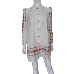 May include: A white long-sleeved tunic dress featuring a pointed collar and a front button-down closure with bright orange buttons. The sleeves and the bottom hem are decorated with intricate, colorful floral embroidery in red, orange, and black.
