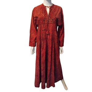Puede incluir: Un vestido largo rojo con un patrón detallado. El vestido tiene mangas largas y un lazo en el escote. La tela parece ser un material texturizado, y el vestido se exhibe en un maniquí.