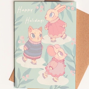 May include: A greeting card with the words "Happy Holidays" and illustrations of a cat, rabbit, and mouse ice skating. The characters are wearing sweaters and dresses in shades of pink and blue. The background is light blue with floral accents.