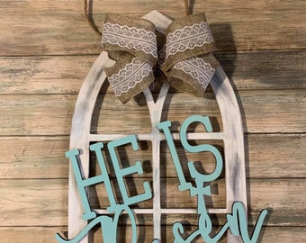 May include: A wooden Easter decoration with a white arched window frame and the words "HE IS Risen" in light blue. It has a burlap bow with white lace trim and a jute rope hanger. The background is a wooden plank wall.
