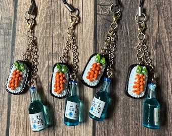 May include: Four miniature sushi charms with a small bottle of soju attached to each. The sushi is made of polymer clay and features a black base, white rice, orange fish eggs, and a green garnish. The soju bottles are blue with white labels and Korean text.