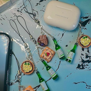 May include: A collection of decorative phone charms featuring miniature food and drink items. Charms include miniature green soju bottles, miniature tarts, and miniature chocolate bars. The charms are attached to black lanyards and silver-colored chains.
