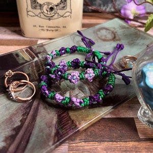 May include: Three purple and green friendship bracelets with small purple flower accents. The bracelets are on a glass tray with two gold rings.