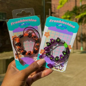 May include: Two colorful friendship bracelets with flower charms on a white and purple background. The bracelets are packaged in clear plastic with a FrankieeMoo logo.