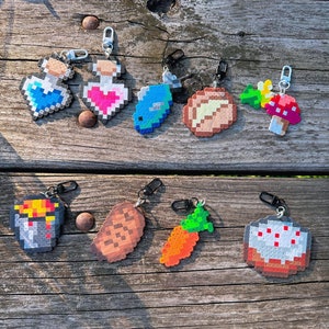 May include: A set of 10 pixel art keychains featuring various designs, including a heart-shaped potion, a fish, a loaf of bread, a mushroom, a bucket, a potato, a carrot, and a cake. The keychains are made of colourful plastic beads and have a silver metal ring.