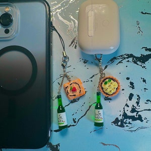 May include: A smartphone and wireless earbuds with decorative charms. The charms include miniature green soju bottles and food items like a tart and a piece of toast with a strawberry.
