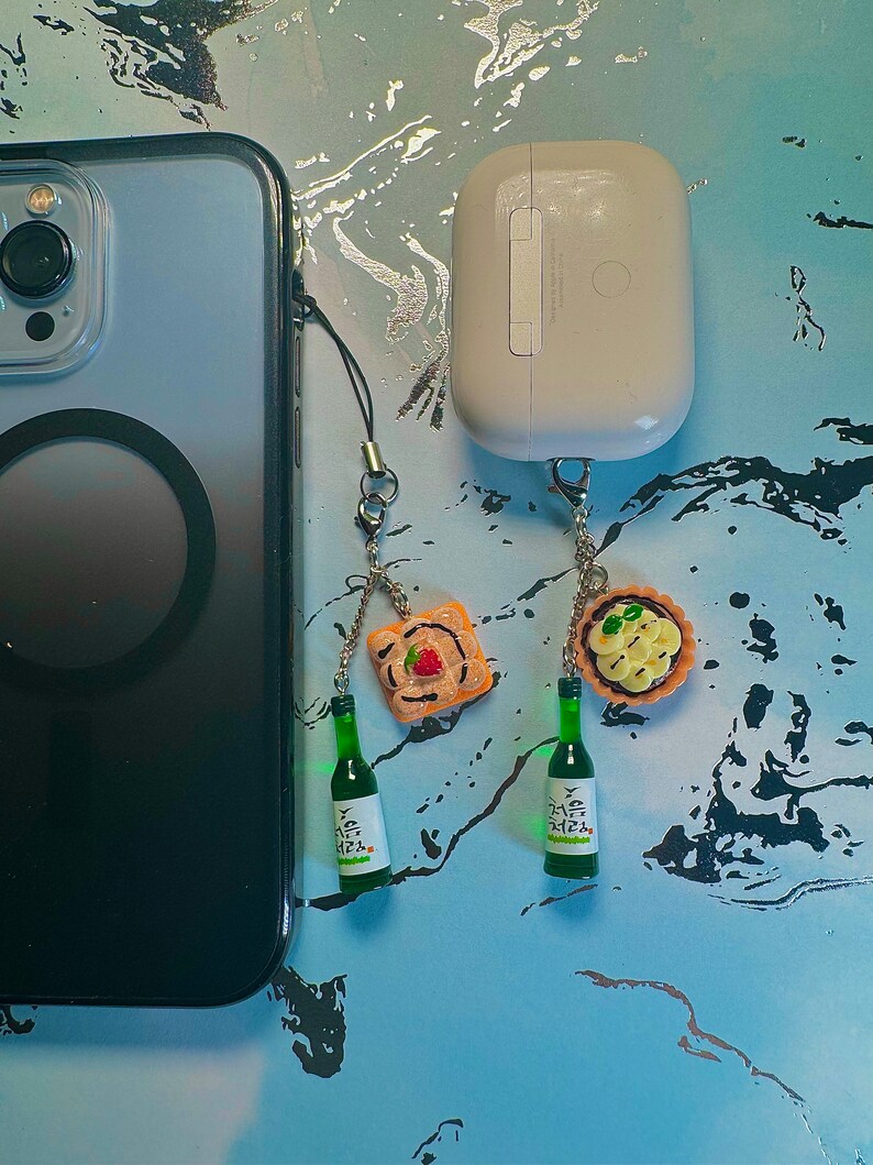 May include: A smartphone and wireless earbuds with decorative charms. The charms include miniature green soju bottles and food items like a tart and a piece of toast with a strawberry.