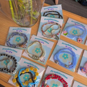 Puede incluir: Una colección de pulseras de cuentas en varios colores y diseños, exhibidas en tarjetas con el logo "FrankieeMoo". Las pulseras incluyen cuentas negras, azules, rojas y doradas, algunas con dijes y detalles de caritas sonrientes. Las pulseras están sobre una superficie de madera.