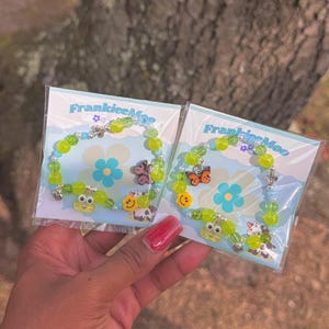 May include: Two green and white beaded bracelets with charms. The charms include a frog, a cow, a butterfly, and a flower. The bracelets are packaged on cards with the text "FrankieeMon".