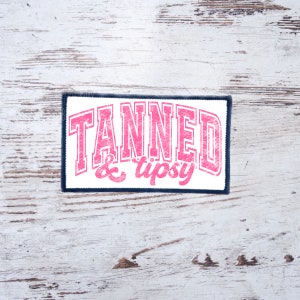 May include: A white patch with a navy blue border featuring the text "TANNED & tipsy" in pink lettering.