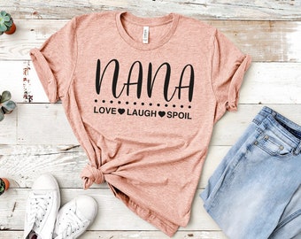 May include: A pink t-shirt with the text "NANA" in black script lettering. Below the text are the words "LOVE LAUGH SPOIL" in black lettering with a heart symbol between each word.