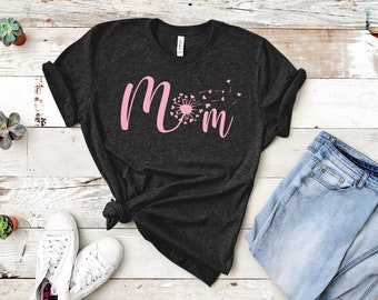 May include: A dark grey t-shirt with the word "Mom" in pink lettering. The "O" in Mom is a pink dandelion with pink hearts blowing in the wind.