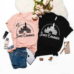 May include: Two t-shirts with a black and white graphic of Cinderella's castle with the text "Just Engaged" on each shirt. One shirt is pink and the other is black.