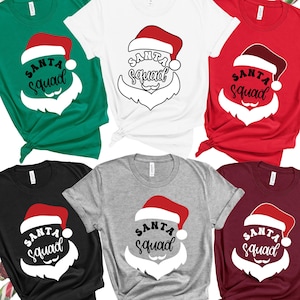 Puede incluir: Seis camisetas de diferentes colores con un gráfico de Santa Claus y el texto "Santa Squad" impreso en la parte delantera. Las camisetas son de color verde, blanco, rojo, negro, gris y burdeos.