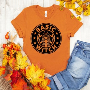 Könnte beinhalten: Orangefarbenes T-Shirt mit einer schwarzen Grafik einer Hexe mit Hexenhut in einem Kaffeetassenlogo. Der Text auf dem Logo lautet "Basic Witch".