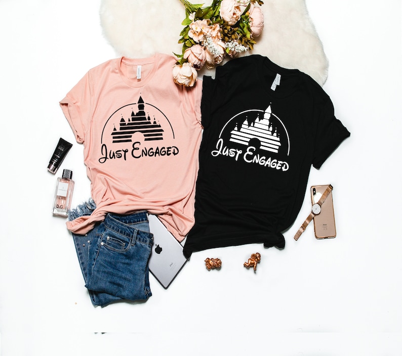 May include: Two t-shirts with a black silhouette of Cinderella's castle and the text "Just Engaged" in white on a black shirt and black on a pink shirt.