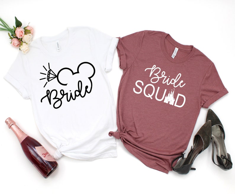 Peut inclure: Deux t-shirts blancs avec du texte noir. Le t-shirt de gauche a une bague de fian&ccedil;ailles et des oreilles de Mickey Mouse avec le mot "Bride" en dessous. Le t-shirt de droite est marron avec du texte blanc qui dit "Bride Squad" avec un graphique de ch&acirc;teau blanc.