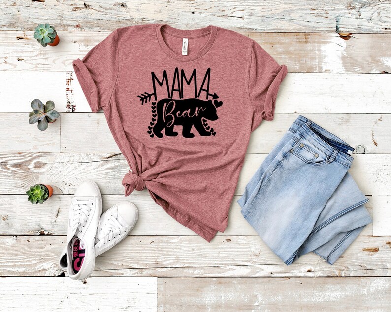 May include: A mauve t-shirt with a black graphic of a bear with the text "Mama Bear" and an arrow pointing to the right. The bear is surrounded by small hearts.