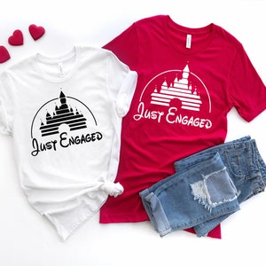 May include: Two white t-shirts with a black silhouette of Cinderella's castle and the text "Just Engaged" on each shirt. One shirt is red and the other is white. A pair of blue jeans is also in the image.