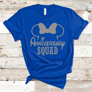 May include: Royal blue t-shirt with a grey graphic. The graphic features Mickey Mouse ears with a bow and the words "Anniversary Squad" in a playful font. The shirt is knotted at the bottom.