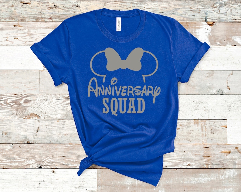 May include: Royal blue t-shirt with a grey graphic. The graphic features Mickey Mouse ears with a bow and the words "Anniversary Squad" in a playful font. The shirt is knotted at the bottom.