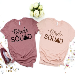 Peut inclure: Deux t-shirts avec le texte "Bride Squad" et une silhouette du ch&acirc;teau de Cendrillon. Les chemises sont dans des tons de rose et de mauve.